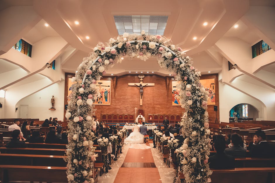 A wedding ceremony inside a church, featuring a large floral arch composed of white and pale pink roses, white baby's breath, and greenery at the entrance. The bouquet arrangement is lush and rounded,