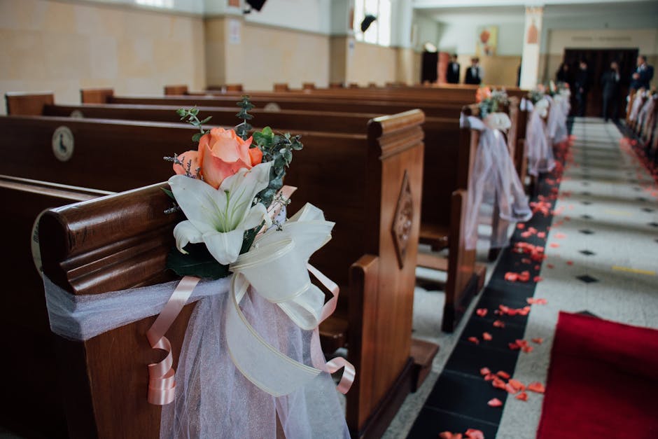 A close-up of a wooden church pew decorated with a floral arrangement on the armrest, featuring a peach-colored rose, white lilies, green foliage, and small purple accent flowers, all wrapped in white