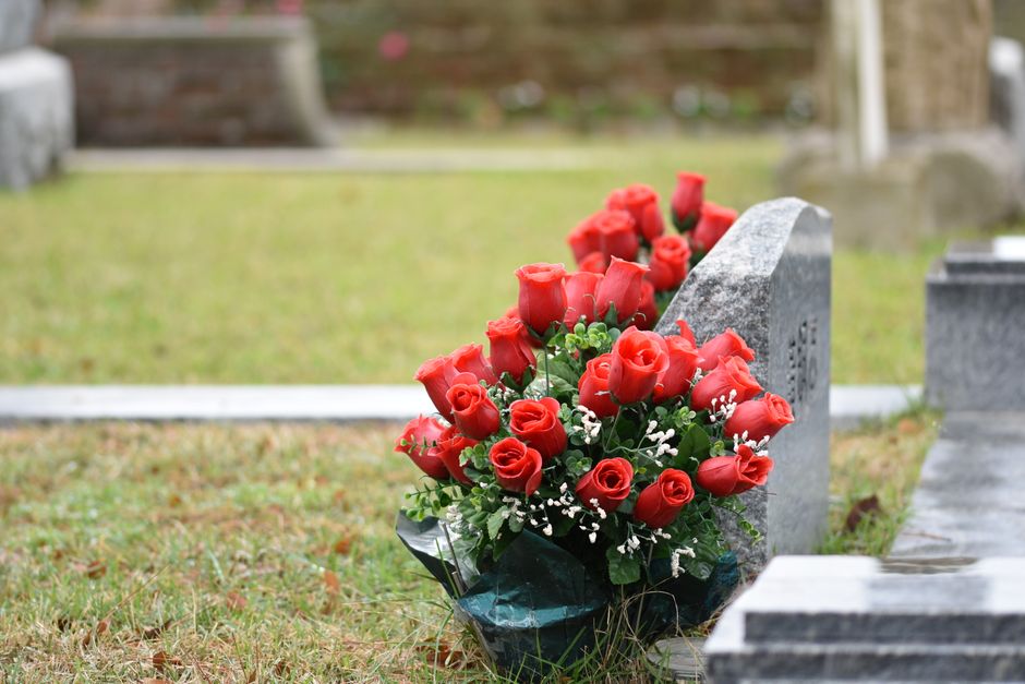 A bouquet arrangement of fresh red roses with green foliage and small white filler flowers, wrapped in dark green and black floral paper, rests against a grey stone memorial plaque on a grassy area. T