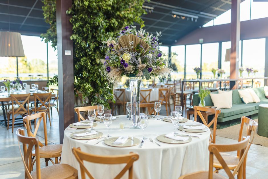 A floral arrangement featuring a large bouquet of white, purple, and cream-colored flowers, including roses, statice, and grasses, is set in a tall, clear glass vase placed at the center of a round di