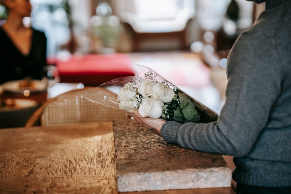 A person is holding a floral gift consisting of white roses and small baby's breath flowers, wrapped in clear cellophane, with a few green leaves visible. The bouquet is being presented over a wooden