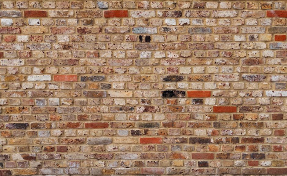 A close-up view of a textured brick wall composed of irregularly arranged bricks in shades of beige, tan, red, brown, and some dark patches. The bricks have a weathered appearance with visible mortar