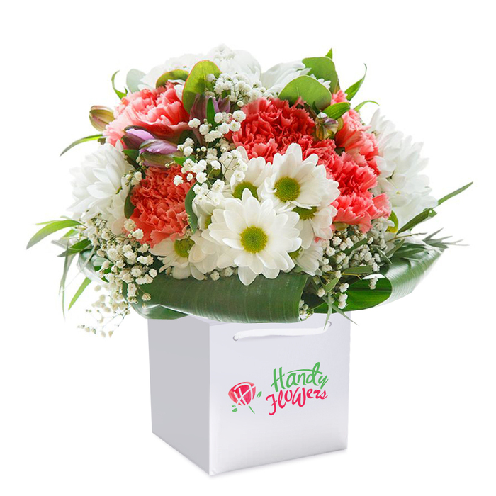 Prepared by skilled hands, this lush bouquet stands tall in a flawless, square white gift bag, offering a graceful nod to the vibrant community of Harrow Weald. The arrangement is anchored by plush coral, peach, and rich red carnations, their textured, frilled petals brimming with colour and evoking a feeling of festive abundance. Between these primary flowers, snowy white daisies catch the light, their yellow-green eyes bright and welcoming, each one neatly defined and full of energy. Airy sprays of baby's breath are woven throughout, creating a soft, fairy-tale atmosphere that lightens the composition. From the arrangement's left, closed buds with subtle purple highlights introduce gentle intrigue and anticipation, perhaps echoing the rejuvenating energy felt in Harrow Weald's tranquil wooded corners. The base is encircled by a generous ring of broad, polished green leaves, lending depth and structure, reminiscent of the thriving greenery along Weald Lane. The white gift bag features chic, durable ribbon handles for effortless carrying or delivery. Bathed in professional lighting, set against a strikingly pure white background, every flower and leaf stands out, unmistakably fresh and expertly composed. Perfect for important milestones, tender gestures, or simply to lift a loved one's spirits, this arrangement delivers the best of local floristry across Harrow Weald.